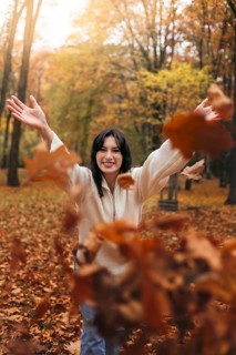 A joyful woman in a cozy sweater, standing amidst falling autumn leaves in a forest. The warm,