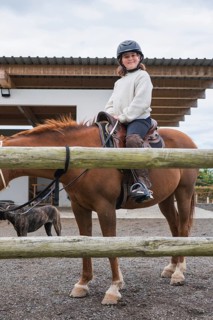A young girl wearing a helmet enjoys a horseback ride at a farm. The scenery includes a stable,