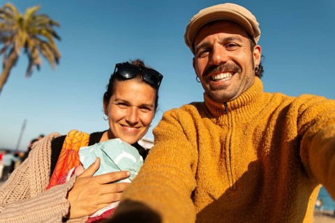 A cheerful couple takes a selfie with their baby on a sunny day in Barcelona. The man wears a
