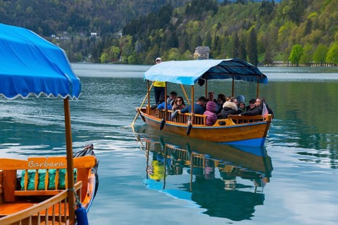 A wooden boat full of people sails on a calm lake surrounded by mountains, Lake Bled, Lake Veldes,