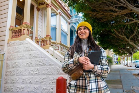 Young woman smiling, holding a tablet while walking on a sidewalk, enjoying urban tourism among