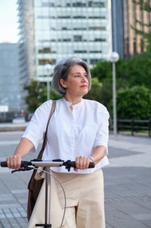 Mature woman with gray hair riding a scooter through a modern cityscape. She looks content and