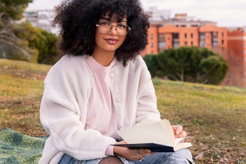 Young latin woman with glasses reads a book while sitting on a blanket in a park. She wears a cozy