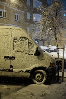 A van becomes blanketed in fresh snow at night, highlighting winter's impact on urban transport in