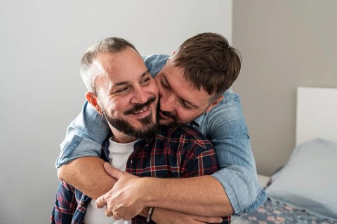 A joyful gay couple embraces lovingly in a cozy bedroom, exuding warmth and affection. One partner