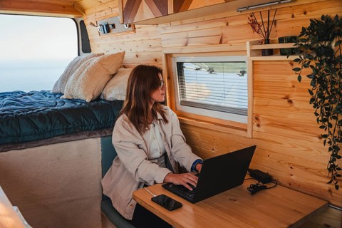 A woman works remotely on her laptop inside a cozy camper van, parked with a scenic view outside.