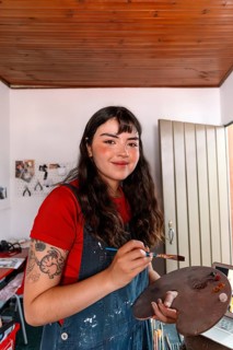 A young woman artist is painting a canvas in her home studio. She holds a palette and brush,