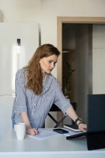 A woman dressed casually in stripes works from home, standing at a table with a laptop, papers, and