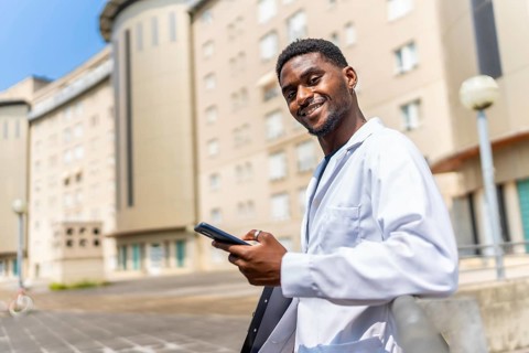African doctor in a work break outside the hospital using mobile and smiling at camera