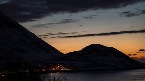 A serene Nordic evening scene captures the silhouette of hills under a crescent moon. The ashen