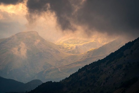 Sunlight pierces through dramatic storm clouds over a mountain landscape, casting a warm glow on