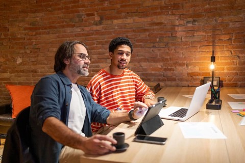 Two professionals in a coworking space engage in discussion, surrounded by laptops and office