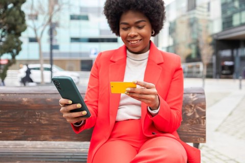 Confident woman in bright red suit sits on a bench, smiling as she uses her smartphone and holding