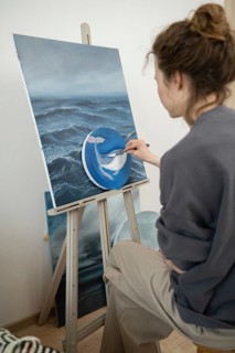 An artist carefully paints an ocean scene on canvas, adding a detailed blue whale. The artwork