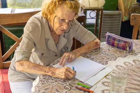 Senior woman sitting at a table with a glass of water and a pencil case, drawing on a notebook with