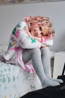 A young girl with curly hair sits on a bed, wearing a colorful sweater and gray leggings. She leans