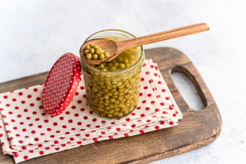A glass jar of fresh green homemade organic peas with a wooden spoon rests on a polka dot cloth on
