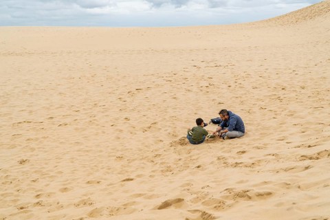 A father and son share a bonding moment, sitting on sand the dune of Pilat, under an overcast sky.