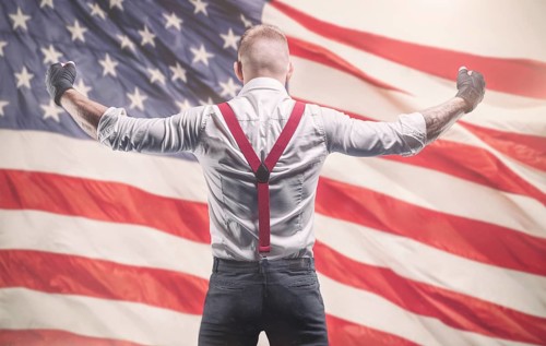 Image of a brutal man in a white shirt and red suspenders standing in front of the American flag.