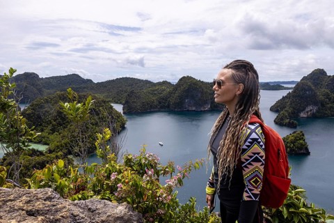 A traveler with braids and a red backpack admires the Raja Ampat archipelago and calm blue waters