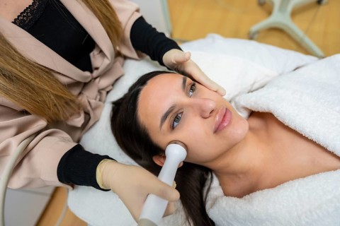 Woman lying on a bed receiving a professional cosmetic facial treatment from a cosmetologist using
