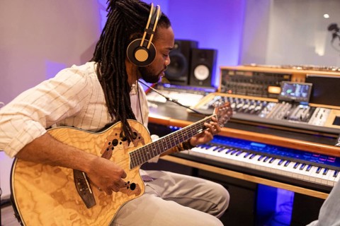 A black musician wearing headphones plays an acoustic guitar in a professional recording studio.