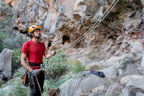 A climber in a red shirt and helmet prepares for traditional climbing on rugged rocks. The natural