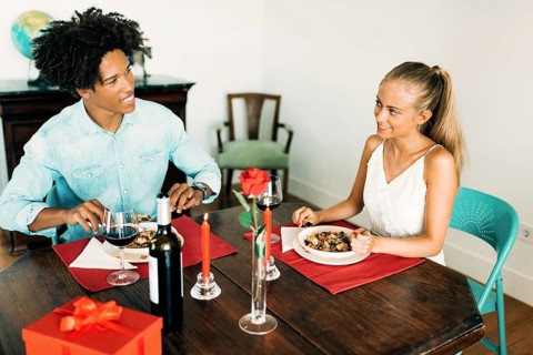 A couple enjoys a romantic dinner indoors, complete with wine, red candles, a rose, and a wrapped