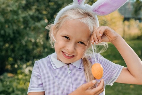 A young child wearing bunny ears joyfully holds a decorated Easter egg in a sunny outdoor setting.