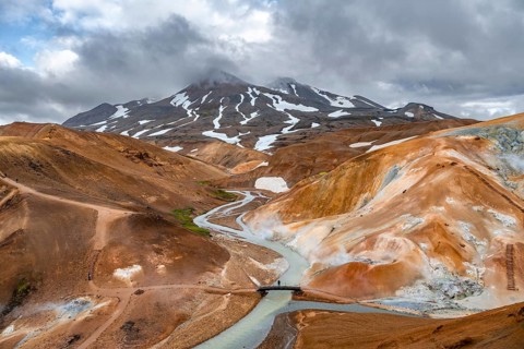 Tourist on bridge over steaming stream between colourful rhyolite mountains with snowfields,