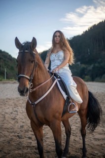 A young woman with long hair rides a brown horse on a sandy beach at sunset. Dressed in white, she