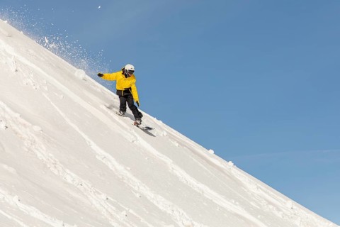 A snowboarder wearing a vibrant yellow jacket carves through fresh powder on a steep mountain