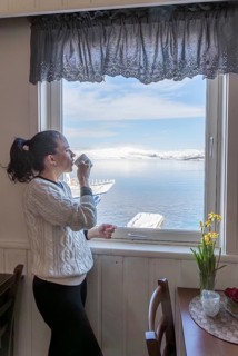 A woman stands by a window, sipping coffee and enjoying a serene view of snow-capped mountains and