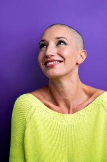A joyful middle-aged woman with a shaved head smiles while looking upward, wearing a vibrant green