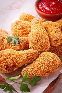 A wooden board holds several golden chicken drumsticks arranged neatly. A small bowl of red dipping