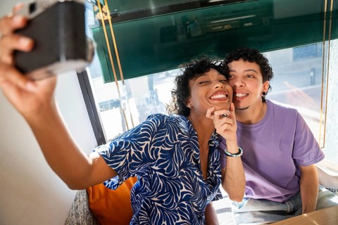 A joyful multiethnic LGBTQ+ couple smiling warmly while capturing a selfie in a cozy cafe. Sunlight