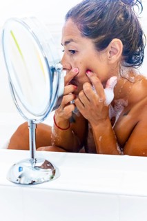 A woman in a bathtub examines her face closely in a round mirror, focusing on skincare. The setting