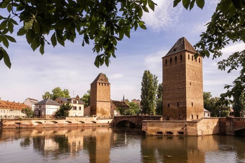 Scenic view of historic towers reflected in the calm waters of the Ill River, surrounded by lush