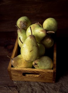 A wooden box is filled with fresh, ripe pears, resting on a burlap cloth. The dark wood background
