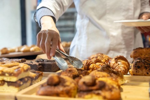 Baker's hand using metal tongs to select a freshly baked croissant from an abundant display of