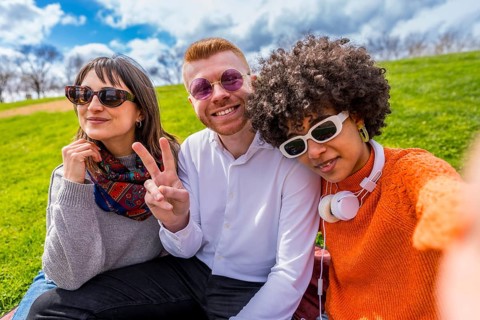 Three young diverse friends are sitting on a blanket in a park, taking a selfie and showing the