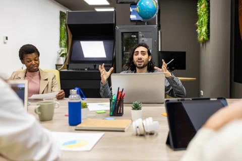 A dynamic office meeting features diverse professionals collaborating around a table with laptops,