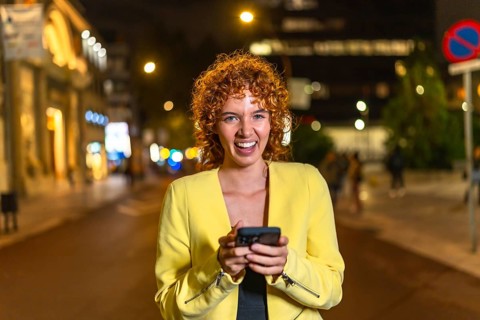 Modern woman in casual clothes and curly hair laughing while using smartphone in the city at night