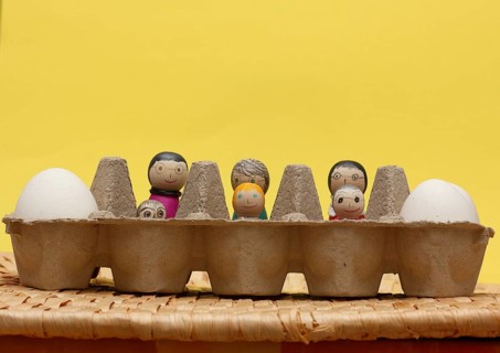 Six amusingly painted wooden cones in an egg carton, against a bright yellow background, North