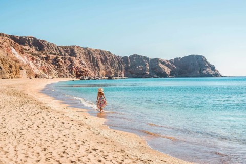 A tranquil scene of a woman in a sundress and hat walking along the sandy shore of Paleochori