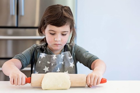 A child in a star patterned apron rolls out dough with concentration. The setting is a bright