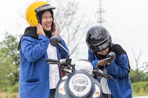 Two Asian Muslim students wearing hijabs and helmets stand with their scooter, ready for a day at