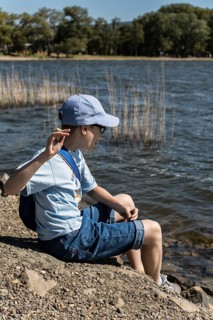 A young boy wearing a blue cap and casual attire sits by a serene lakeside, playfully engaging with