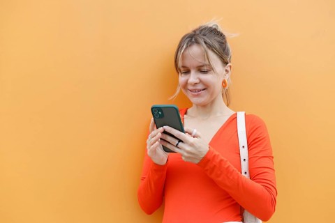 A smiling woman in a red top uses a smartphone against a bright orange wall in Warsaw She is