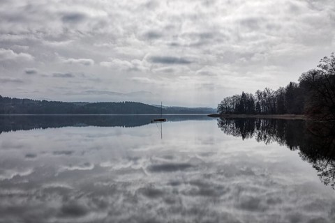 A calm lake with a single boat in the water, surrounded by forest and a cloudy atmosphere, Seehamer